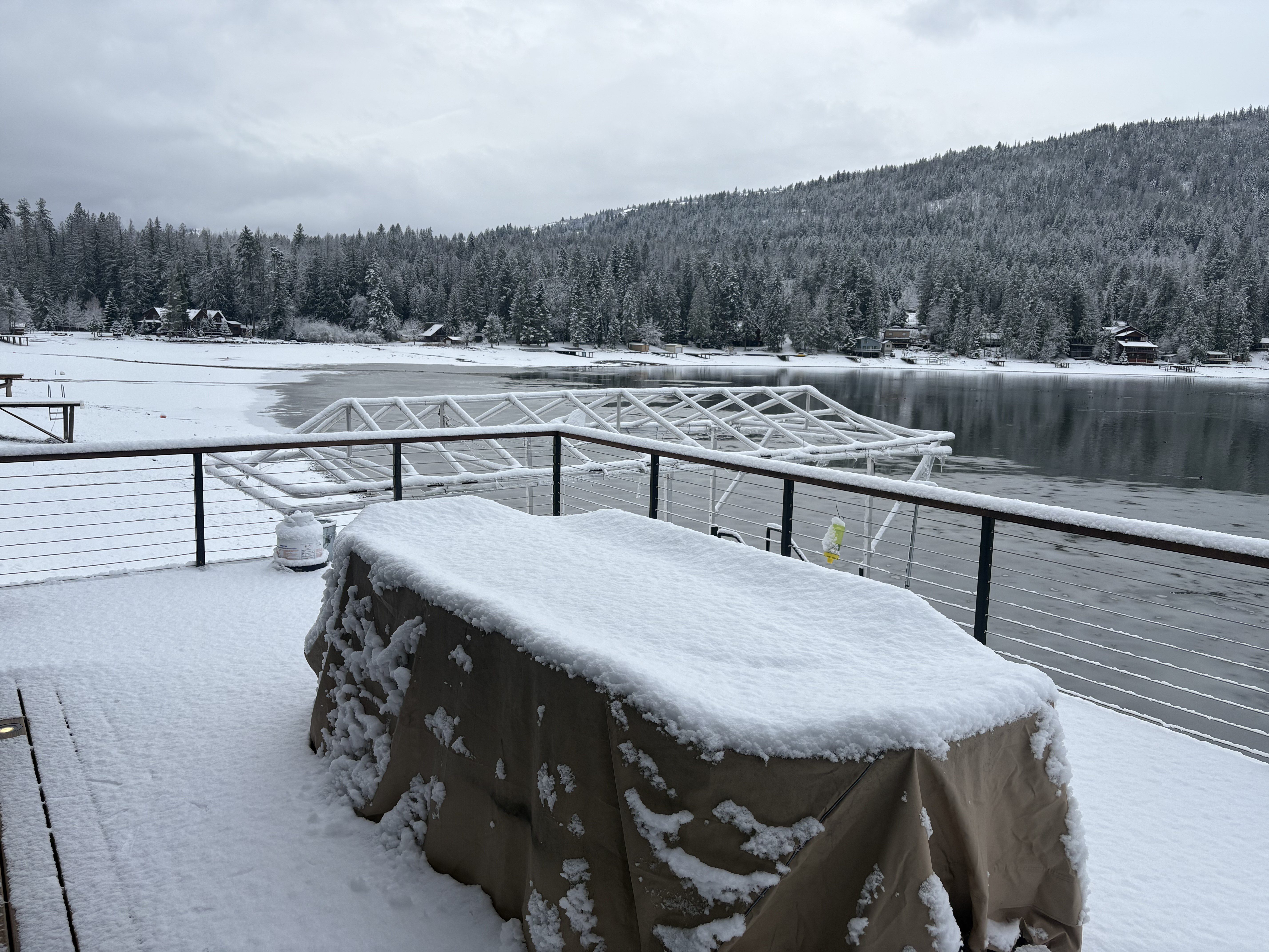 Snow-covered deck overlooking a calm lake with boat lift and forested hills in winter.