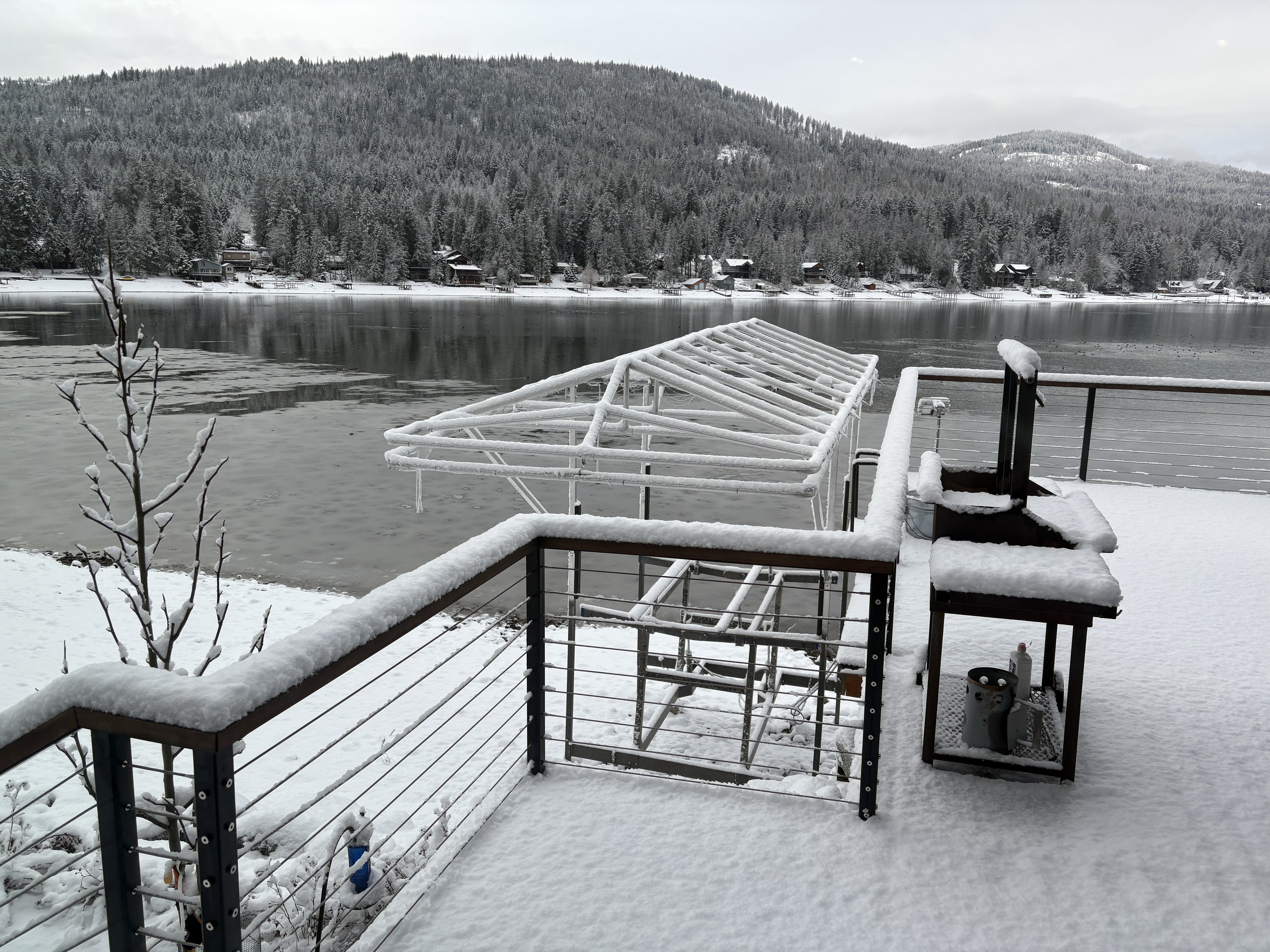 Snow-covered deck with grill overlooking a calm, partially frozen lake and forested hillside.
